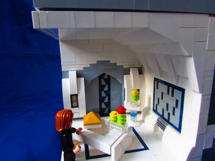 LEGO MOC - Новогодний Кубик 3015 - Земля. Новый 3015 год.: Один из входов ведет в роскошную с высоким потолком и слепящими белизной стенами. По встроенному в стену серому экрану плавно опускаются голубые снежинки. На изящной мебели установлены символы Нового года - необычные растения с яркими цветами венерианские елки, завезенные туда с Земли и приобретшие новые очертания. Они стали очень популярны в современном обществе, полностью вытеснив обычные земные ели из новогодних жилищ. На столе находится сияющая золотом пирамида, Как символ восхождения к высшей форме сущности человека.